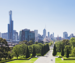 A view of the Melbourne city skyline from the Shrine of Remembrance on a sunny day. In the foreground people walk down a path in the gardens.