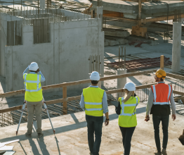 Four construction workers in high-vis vests survey a building site. The building site has new concrete slabs and exposed timber beams. 