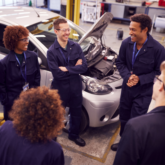 A group of TAFE mechanic students talking and laughing in a classroom workshop.